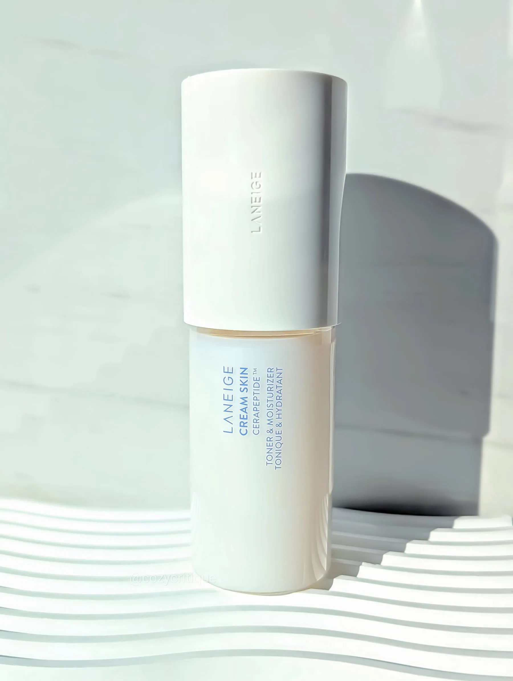 Laneige Cream Skin bottle on white patterned surface, lit by soft sunlight. Text reads "Laneige Cream Skin" and "Toner & Moisturizer."