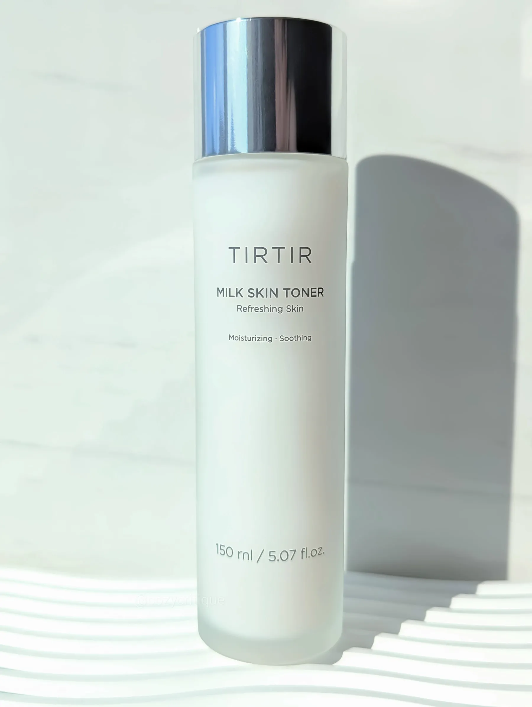 A white bottle of TIRTIR Milk Skin Toner with a silver cap is on a striped surface. Text: "Refreshing Skin, 150 ml / 5.07 fl. oz."