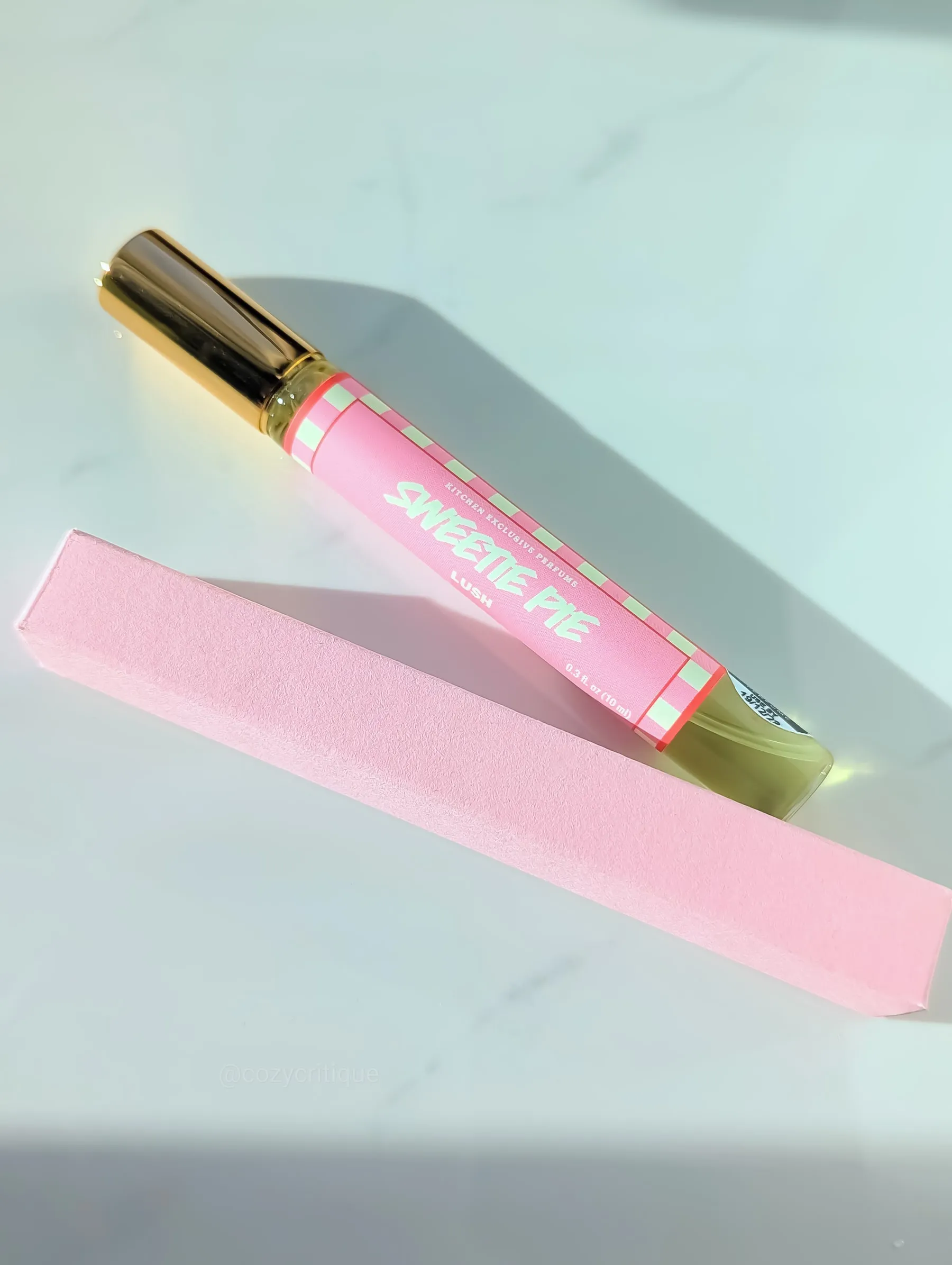 Pink perfume bottle labeled "Sweetie Pie" on marble surface, next to a matching pink box. Golden cap, pastel checkered pattern.