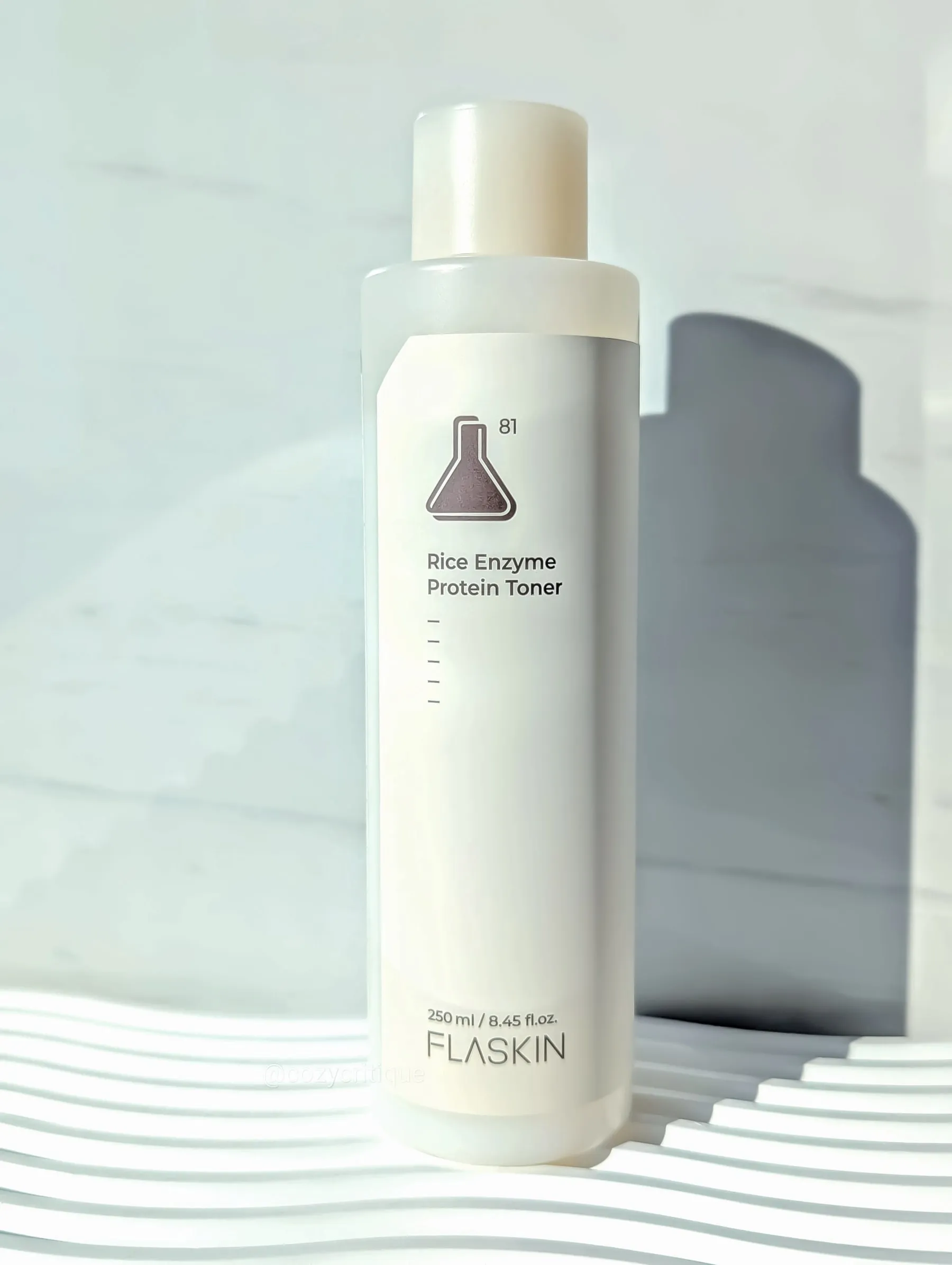 A bottle of Rice Enzyme Protein Toner stands on a wavy, white surface. Labels show "FLASKIN" and "250 ml." Light gray background.