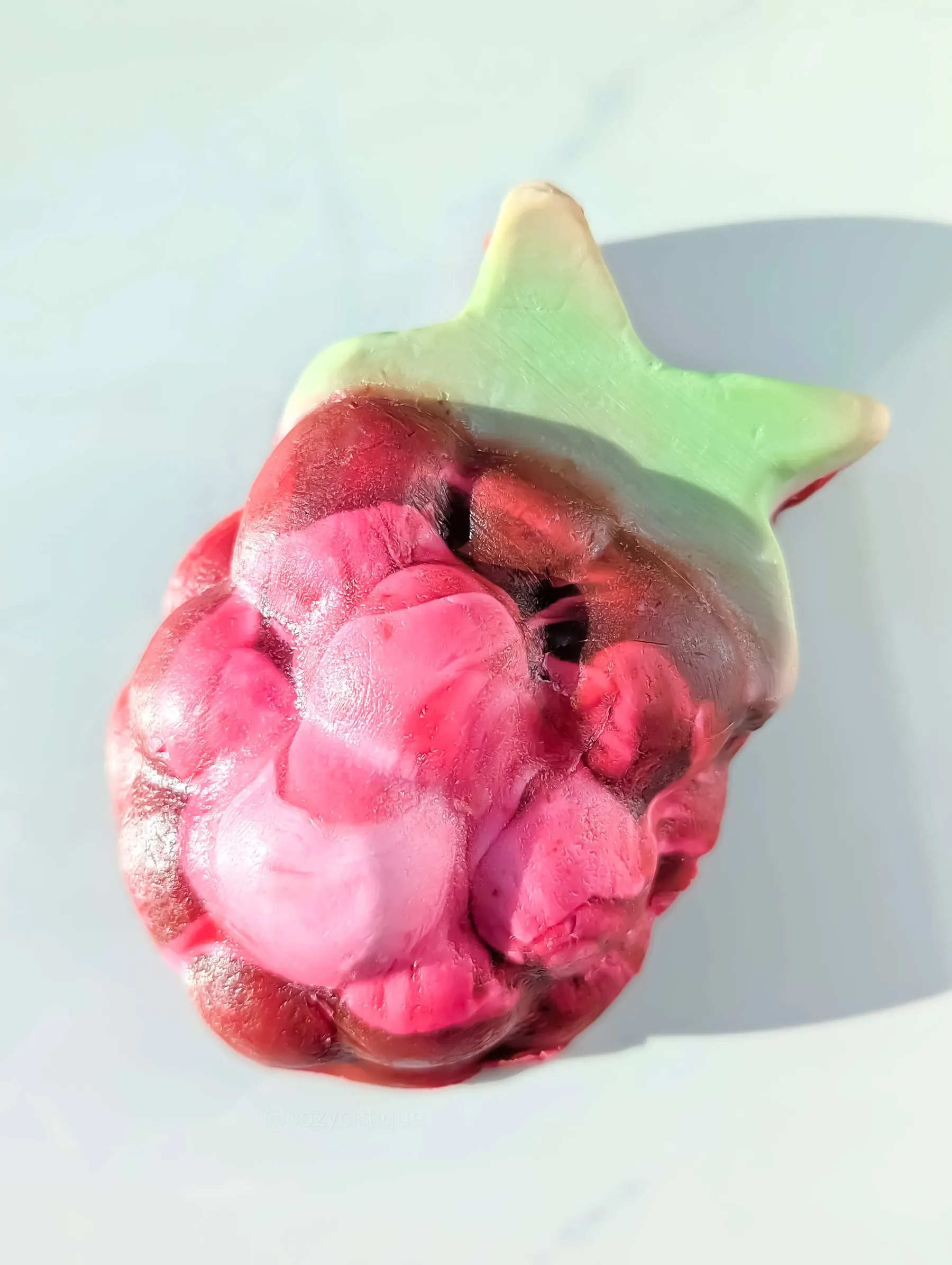 A colorful soap in the shape of a raspberry with green leaf on top, against a light background. Features shades of pink and red.