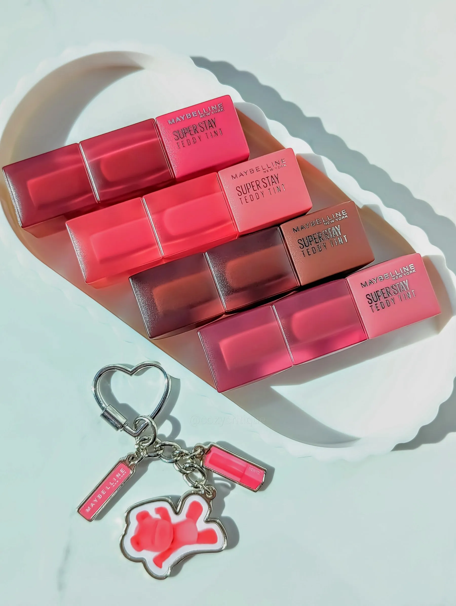 Maybelline lip tints in pink shades on a white scalloped tray. A heart keychain with pink accents and a bear charm is nearby. Bright lighting.