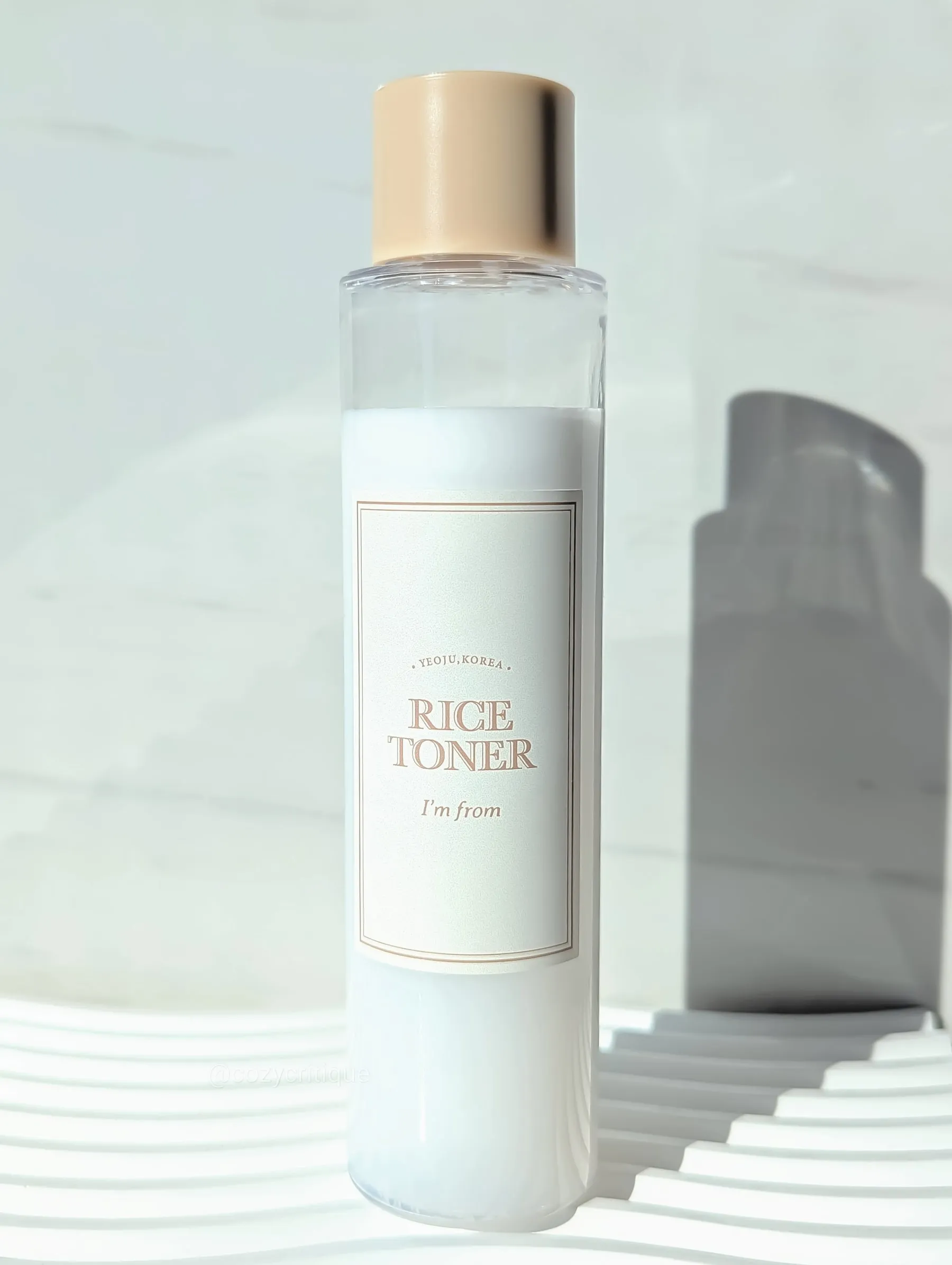 Clear bottle with beige cap labeled "Rice Toner I'm from Yeoju, Korea." Set on white background with soft shadow cast. Minimalist vibe.