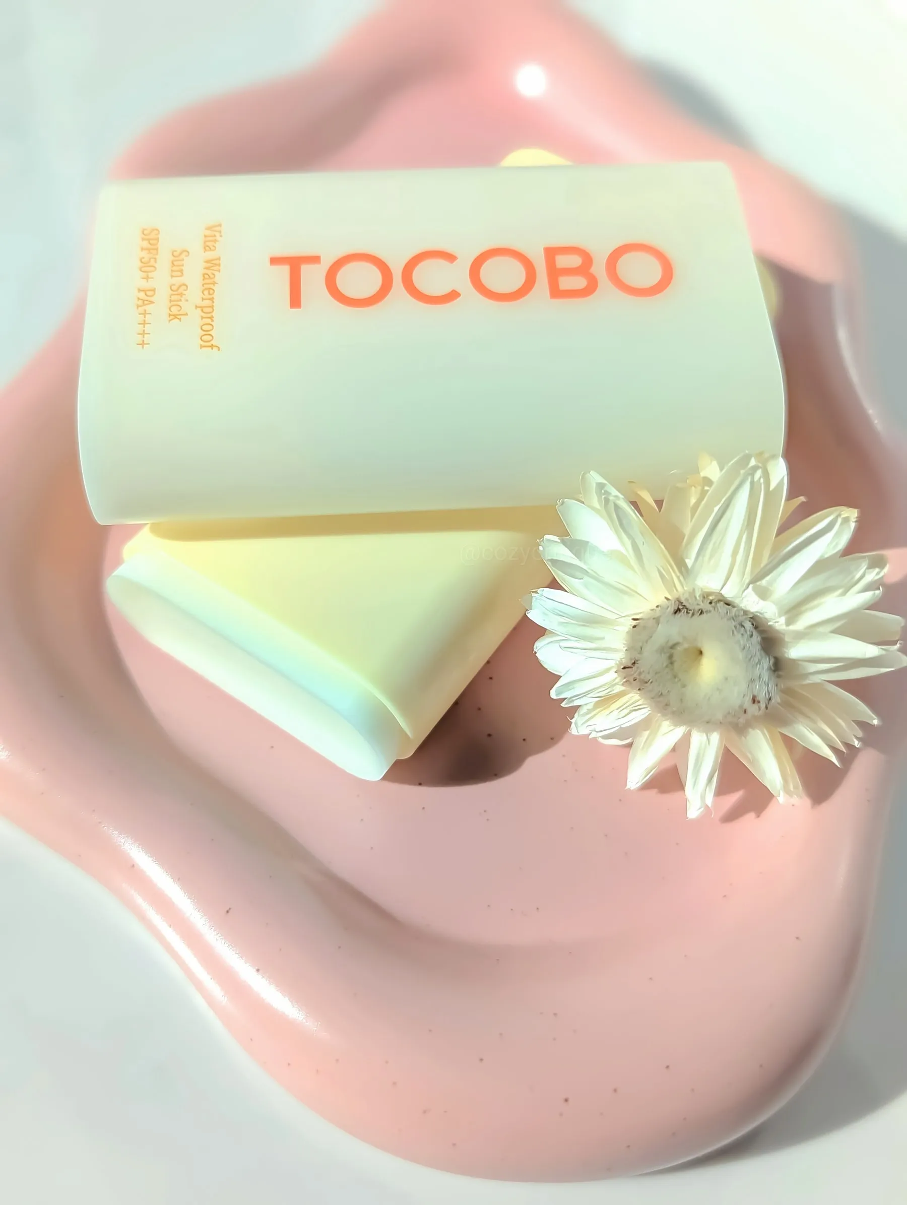 Sunscreen stick labeled TOCOBO on a pink dish with white flower. Text: "Vita Waterproof Sun Stick SPF50+ PA++++". Bright, fresh mood.
