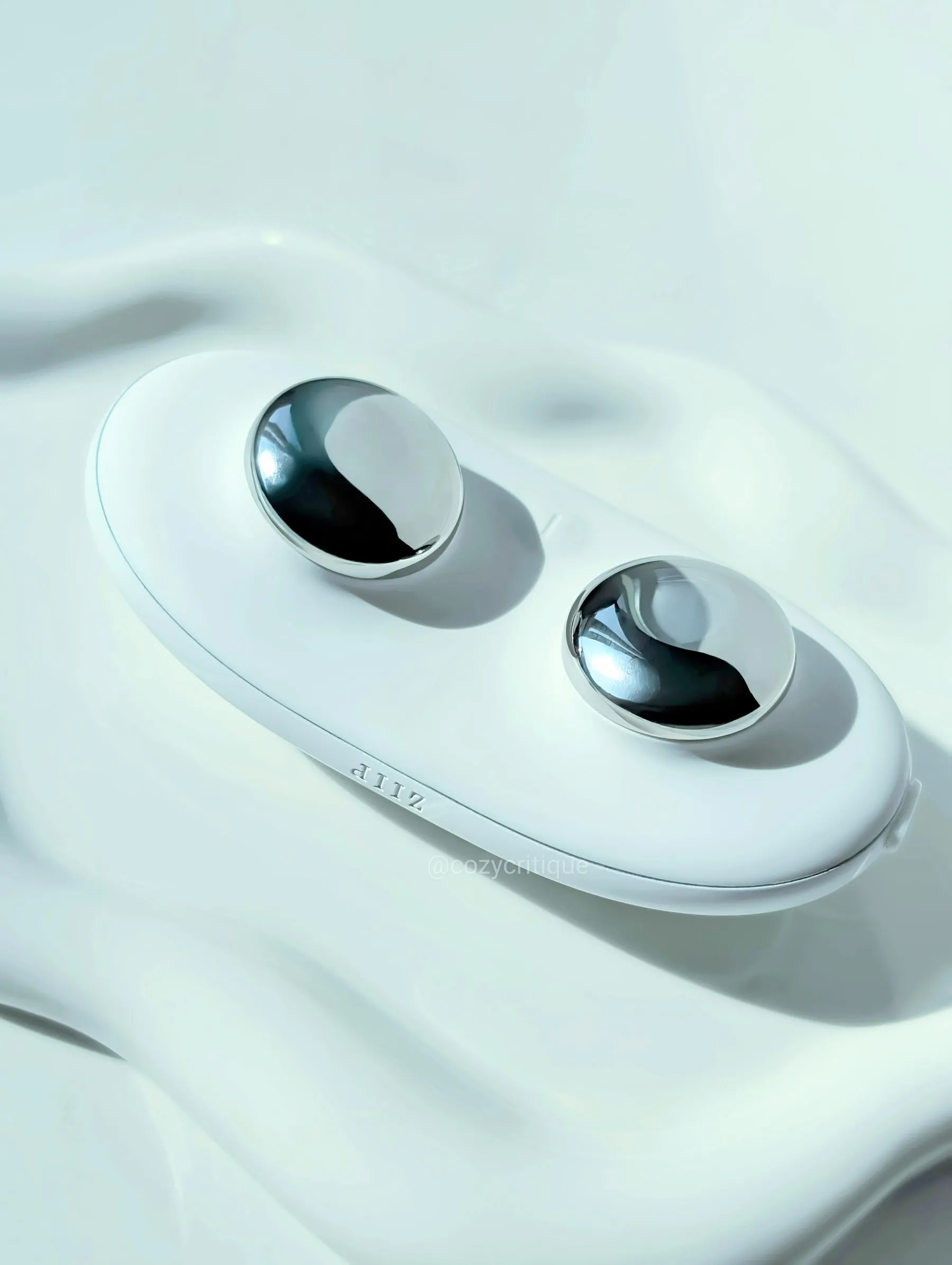 White beauty device with two silver domes on a smooth white surface. Text: “ZIIP” and “@cozycritique” visible. Minimalistic design.