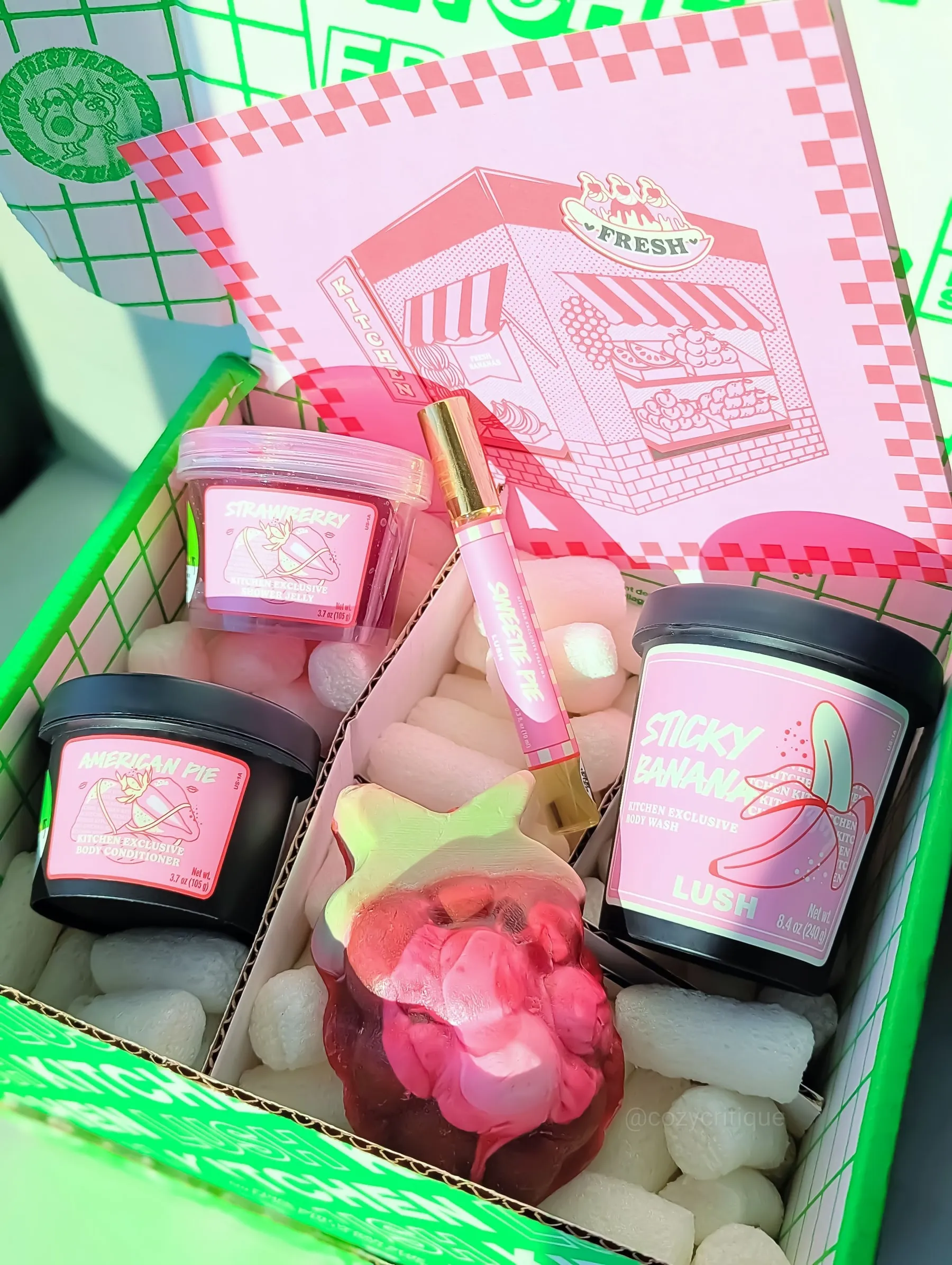Box of Lush products with vibrant pink labels, including body wash and conditioner. Pink leaflet with illustrated storefront. Green box.