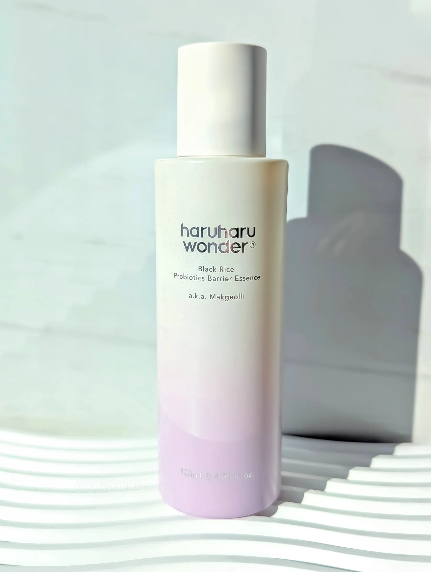 Skincare bottle with text "haruharu wonder" and "Black Rice Probiotics Barrier Essence." Soft gradient on a wavy, pastel-lit surface.