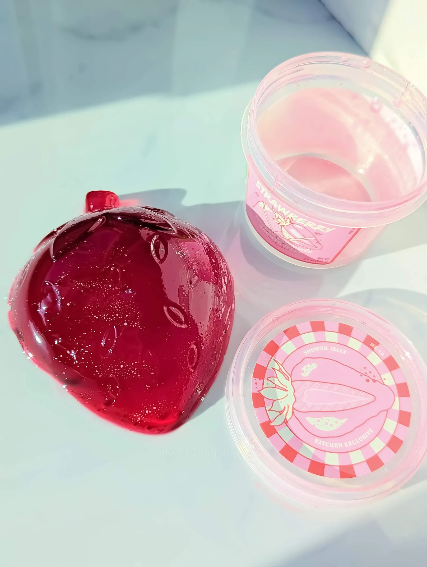 Red jelly shaped like a strawberry and a pink tub labeled "Strawberry Shower Jelly" on a light surface. Bright and playful vibe.